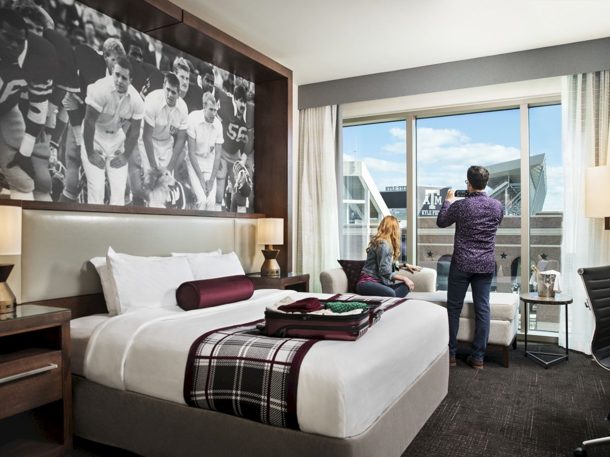 A modern hotel room with a large bed, black-and-white wall art, two lamps, a seating area by the window, and a couple near the window looking outside.