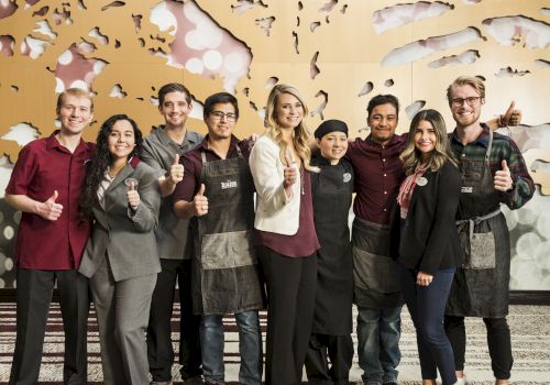 A diverse group of people in aprons posing and giving thumbs up, standing together in a modern indoor setting.