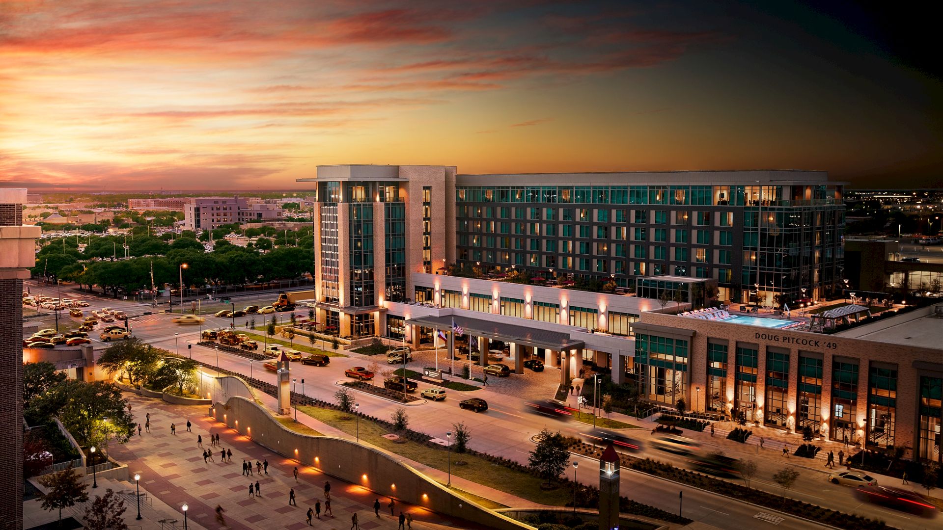 A modern city plaza at sunset: a large glass-front building, wide open square, flowing ramps, and warm orange skies overhead.