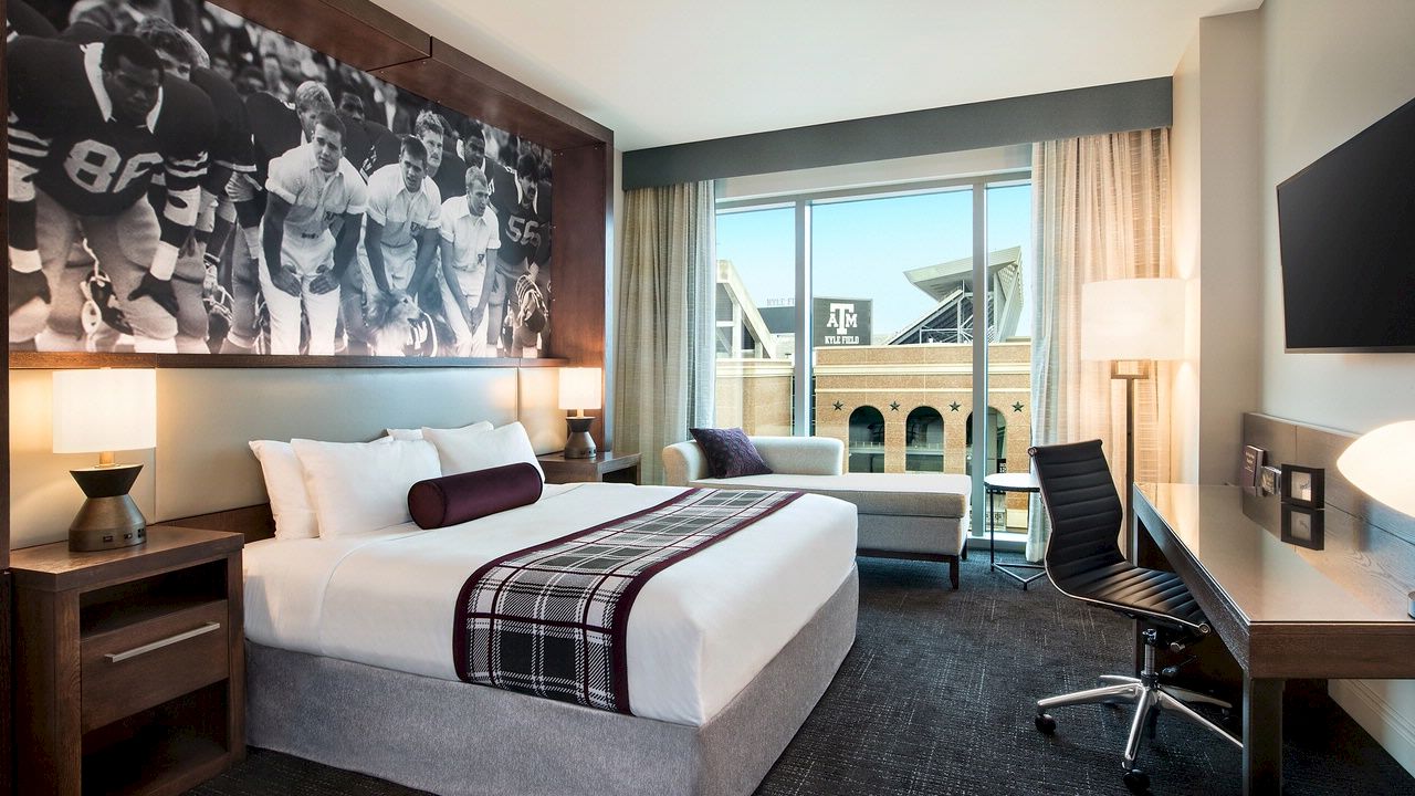 A modern hotel room featuring a tidy bed, Aggie-centric wall art, a desk and office chair, and grey lounging couch overlooking Kyle Field