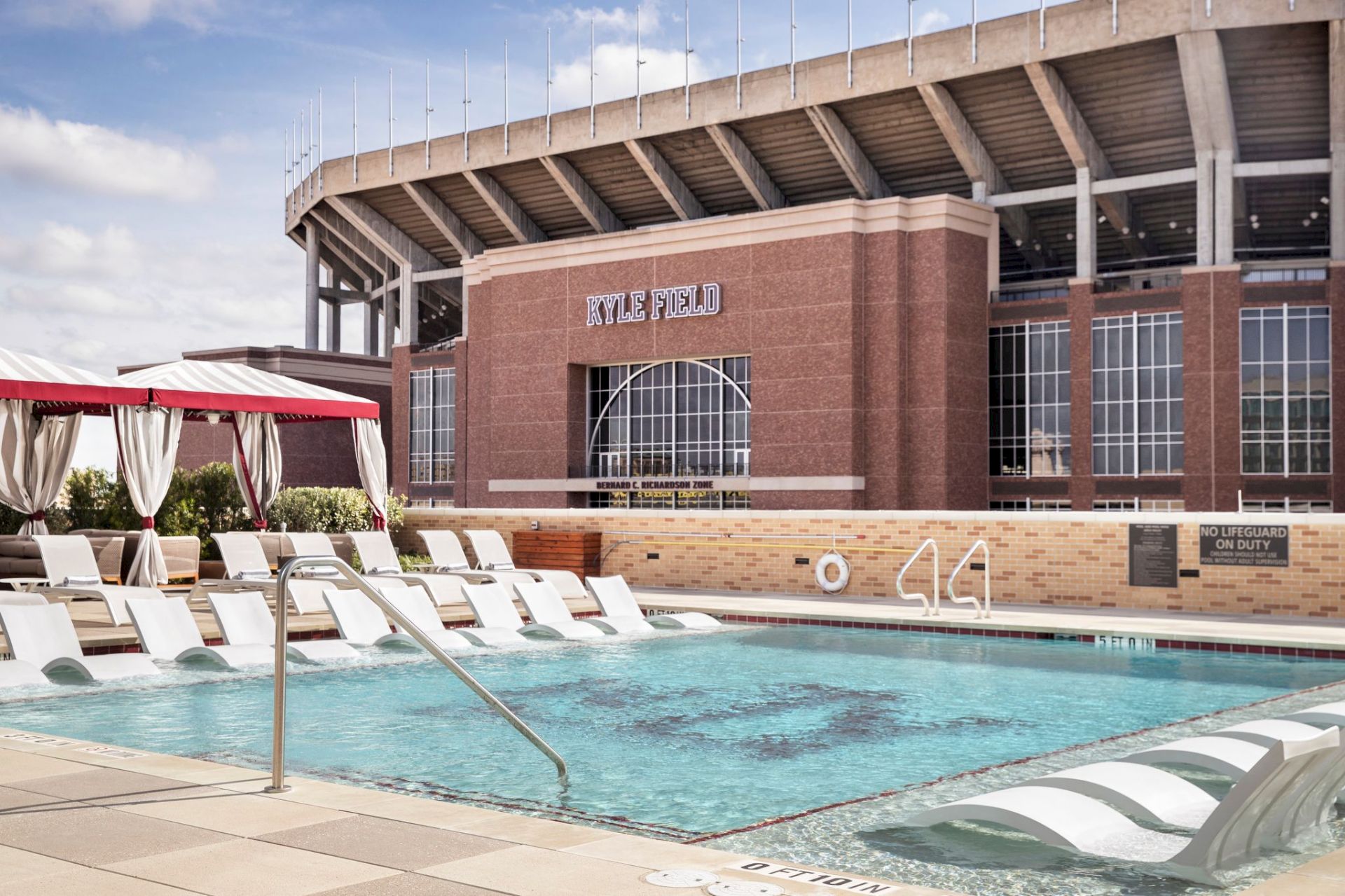 A swimming pool with lounge chairs and a cabana is in front of a stadium labeled "Kyle Field." Sunny day, no lifeguard on duty.