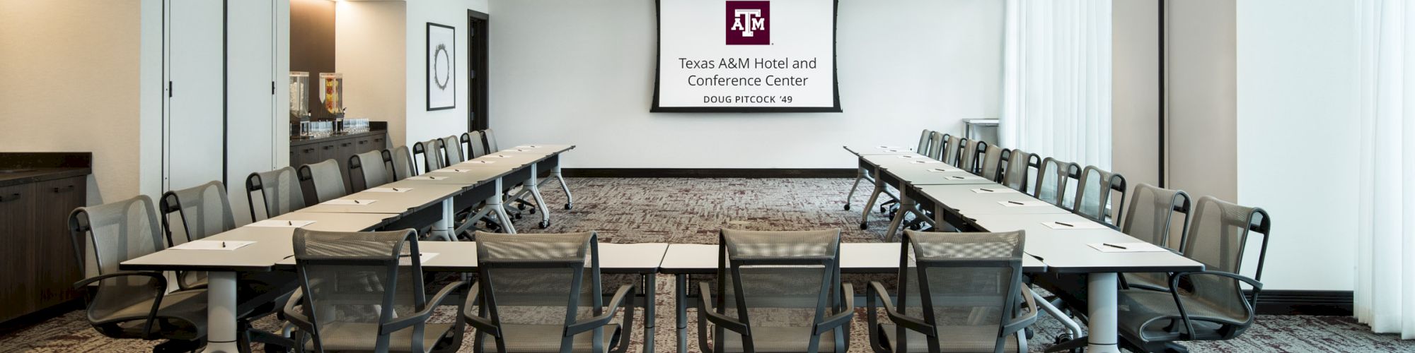 A modern conference room with a U-shaped table setup, multiple chairs, a screen displaying Texas A&M Hotel and Conference Center.