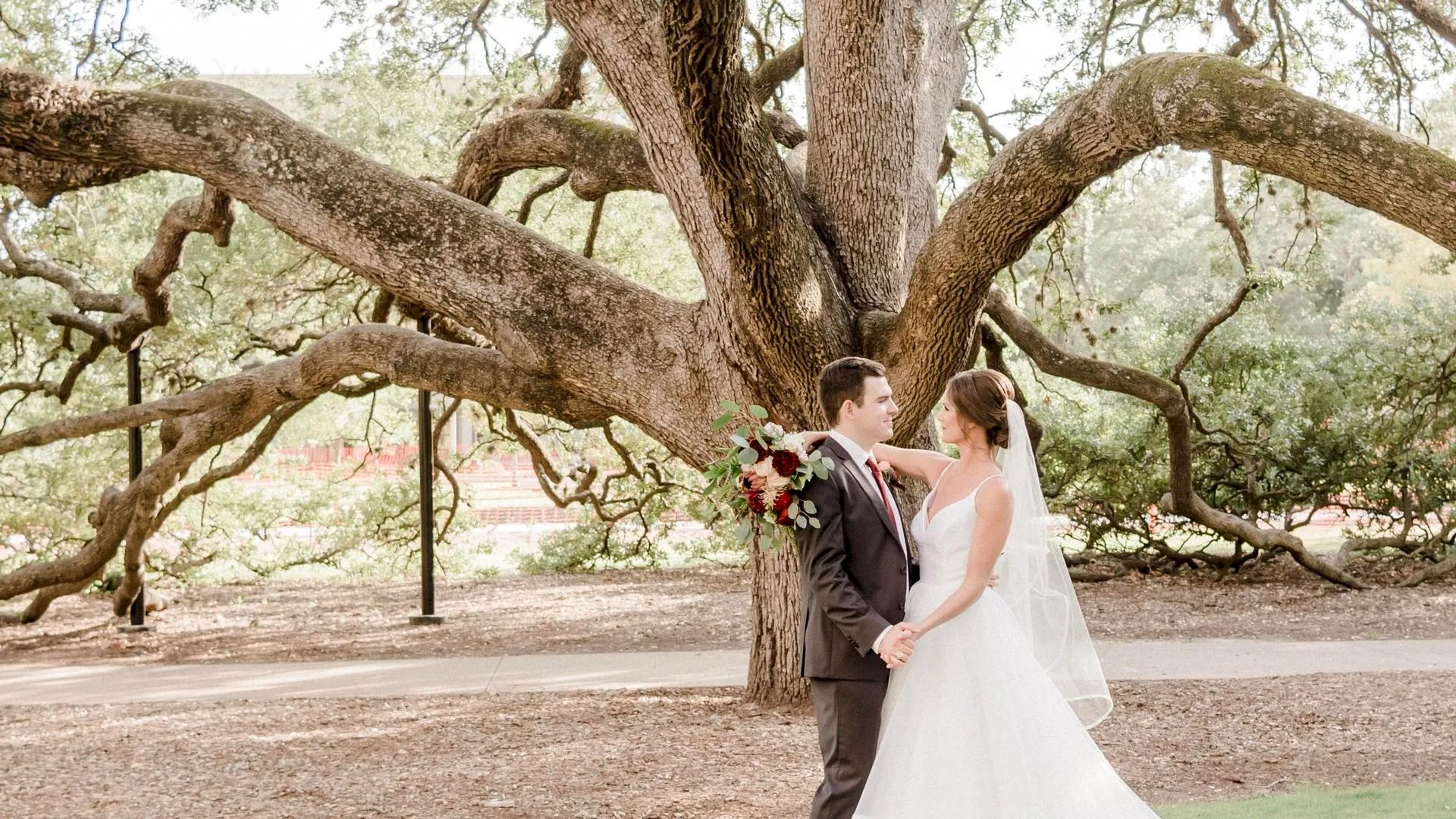 A bride and groom stand under a large, sprawling tree, holding hands and looking at each other, with the bride holding a bouquet of flowers.