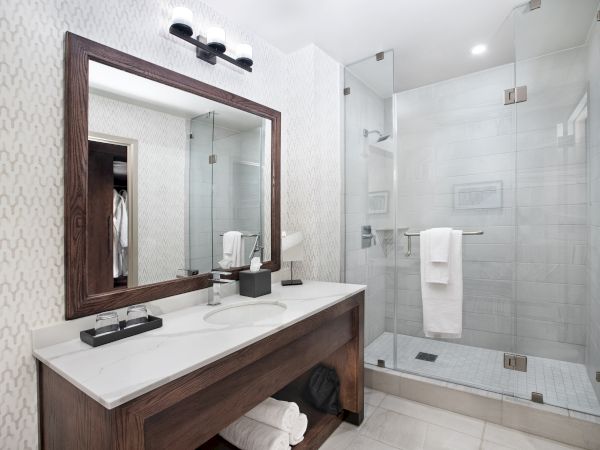 A modern bathroom with a wooden vanity, large mirror, sink, and glass-enclosed shower with towel racks. The decor is minimalist and clean.