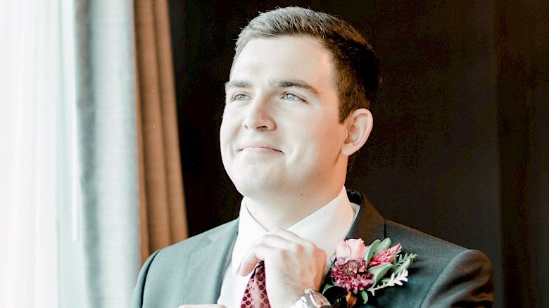 A man in a suit adjusts his tie and stands near a window. He has a floral boutonniere on his jacket.