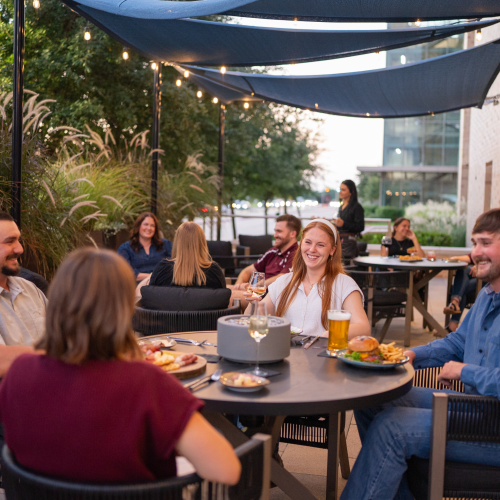 Friends dine and laugh around a patio table under string lights, drinks in hand, in a cozy outdoor gathering.
