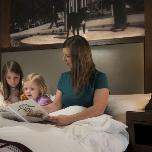 A woman and two children sit on a bed reading a magazine together in a cozy hotel room.