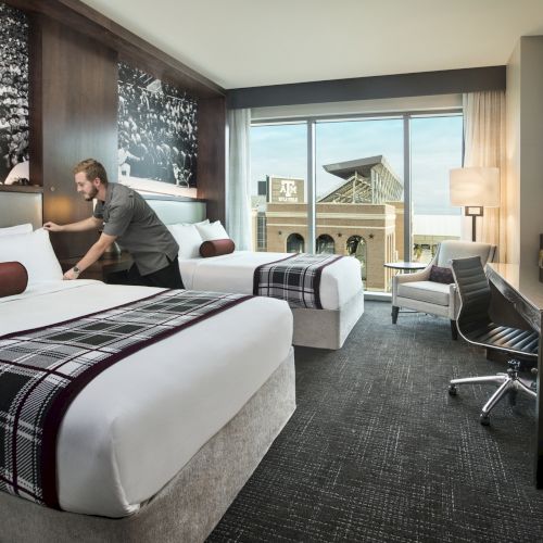 A modern hotel room with two beds, a desk, a chair, a flat-screen, and a balcony view; a guest is tidying the bed, 140 characters.