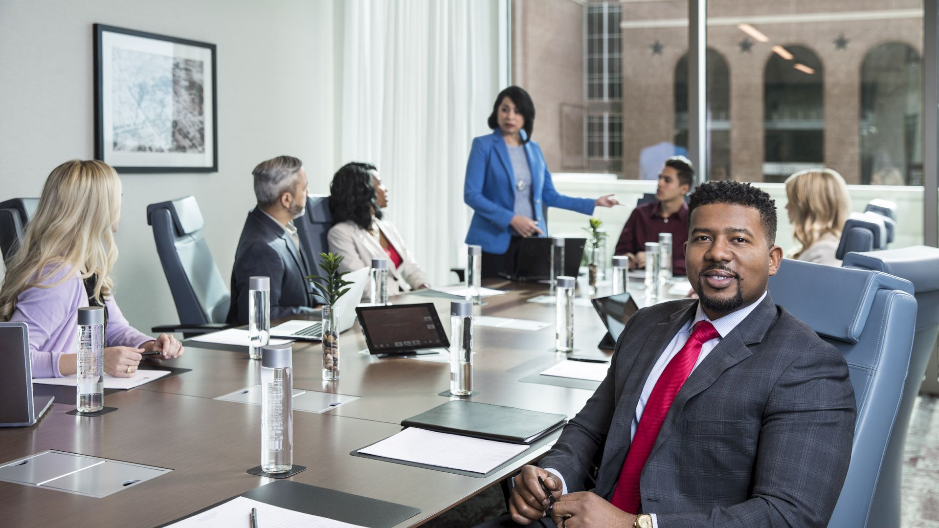 A group of people in business attire are seated at a conference table, with one person standing and speaking, suggesting a meeting or presentation.