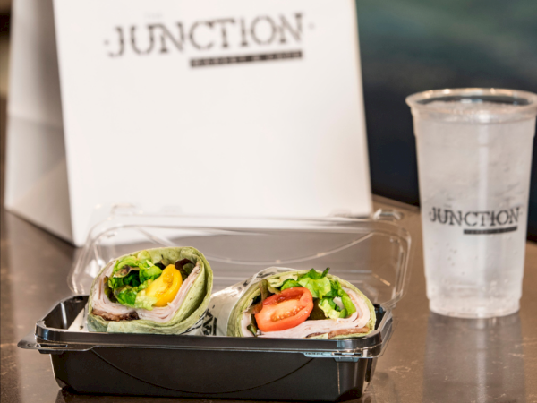 A meal box holds two veggie wraps with tomato and greens, next to a &ldquo;JUNCTION&rdquo; drink cup on a table.