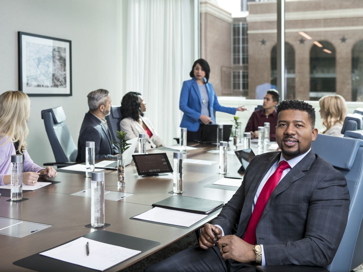 A business meeting in a modern conference room, with a presenter addressing colleagues seated around a long table.