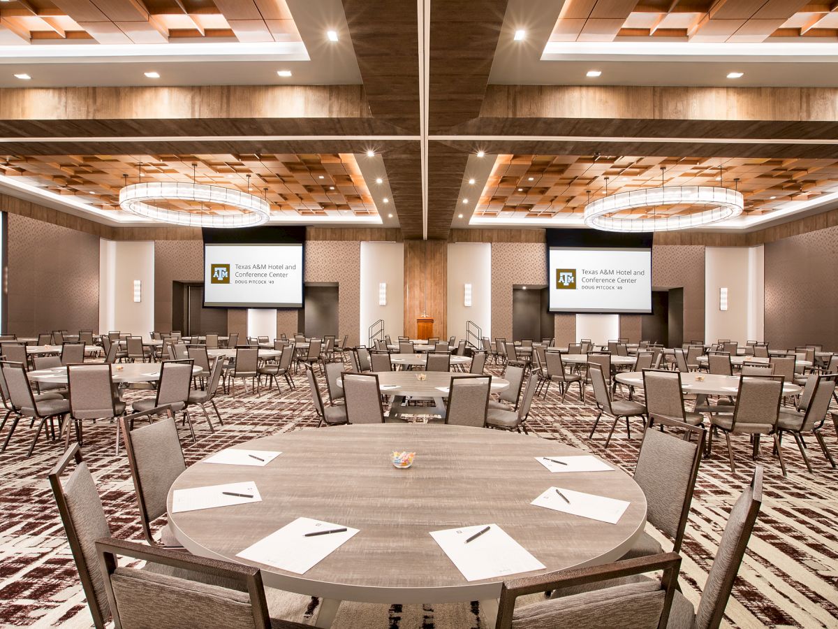 A modern conference room with round tables, chairs, and two projection screens for a Texas A&M Hotel and Conference Center event.