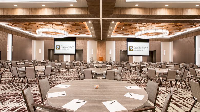 A modern conference room with round tables, chairs, and two projection screens for a Texas A&M Hotel and Conference Center event.