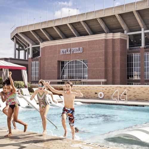 Kids jump into a hotel pool beside lounge chairs; a brick stadium-looking building labeled &ldquo;Nye Field&rdquo; looms in the background, sunny day.