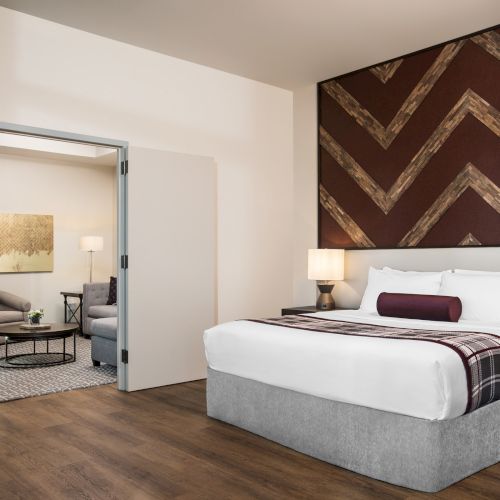 Modern hotel room featuring a tidy bed, sitting area with coffee table, and tv. Above the bed is a rustic wood and maroon art piece