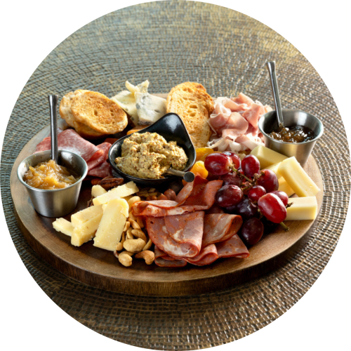 A cheese board with assorted cheeses, cured meats, crackers, nuts, grapes, and dips on a wooden round platter.