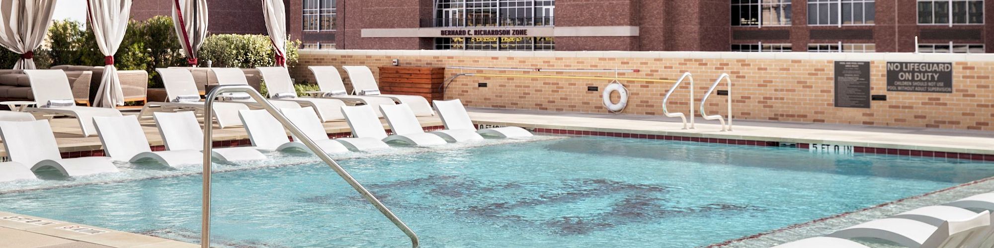An outdoor pool area with lounge chairs and red umbrellas next to a brick stadium or arena building, labeled &ldquo;Metro,&rdquo; on a sunny day.