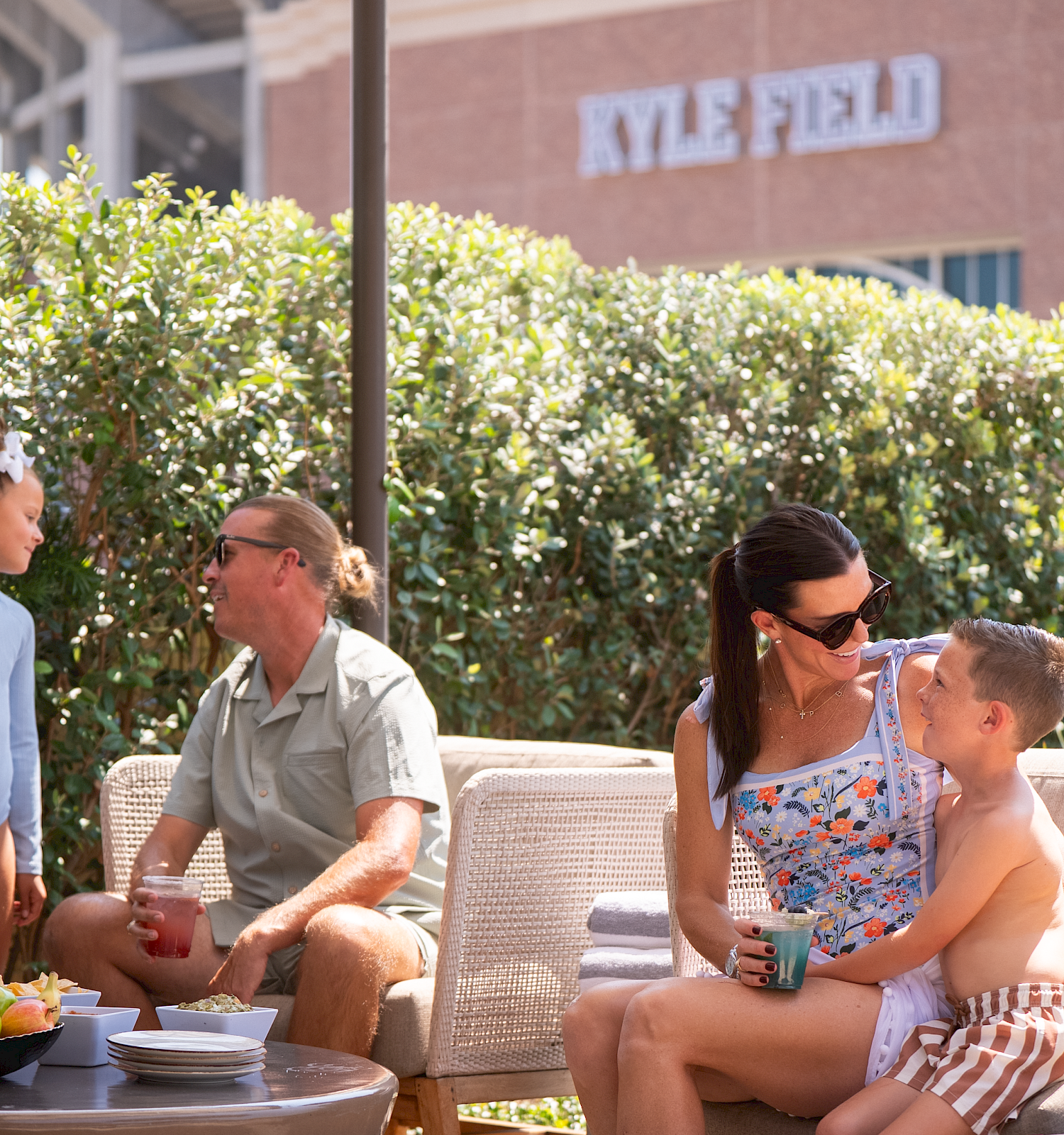 Two couples sit on a patio outside a building, chatting and sharing drinks, with a bowl of fruit on a table and a child cuddled on a mom&rsquo;s lap.