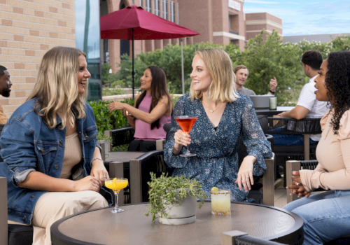 Four friends chat and sip colorful cocktails on a sunny outdoor patio, with umbrellas, brick walls, and greenery in the background.