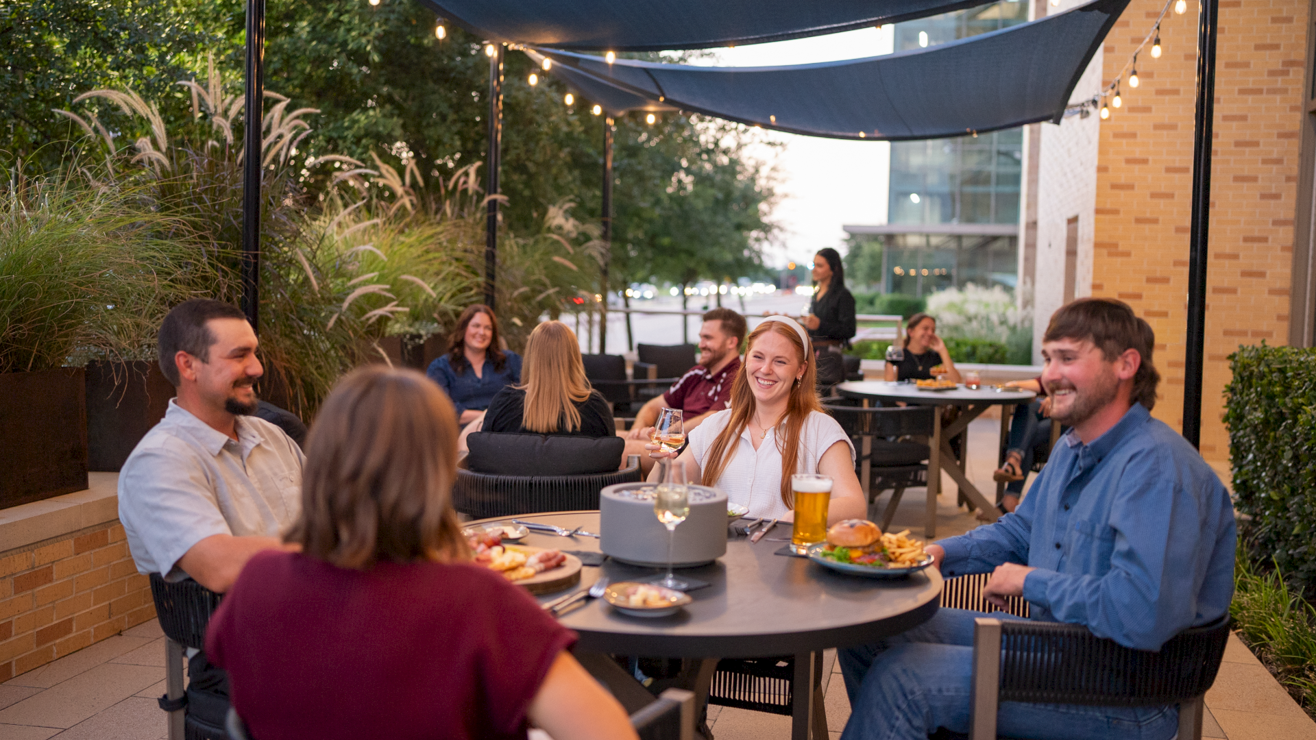A group of people dining outdoors on a patio, chatting and enjoying meals under string lights and shaded canopies.