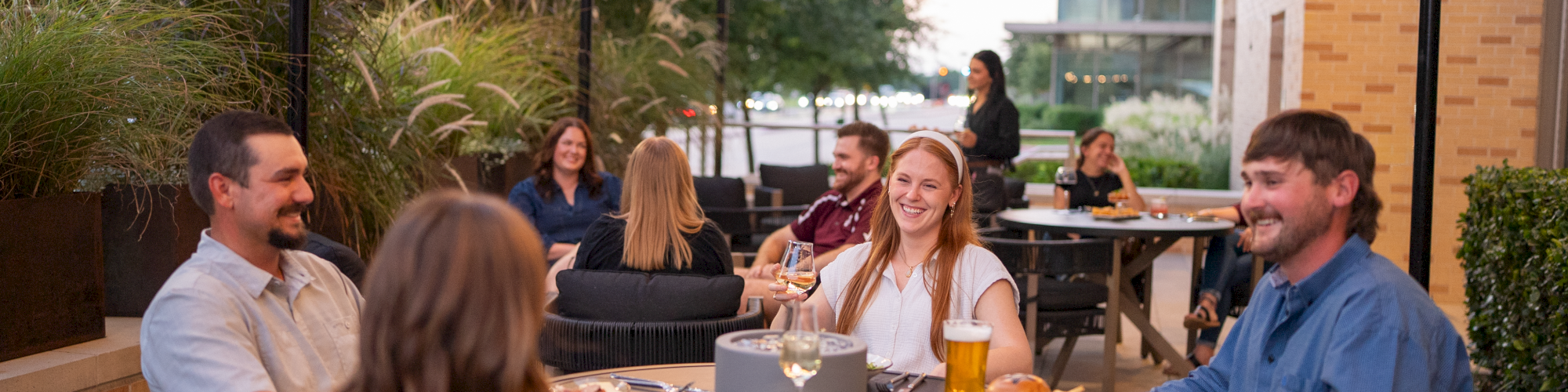 A group of people dining outdoors on a patio, chatting and enjoying meals under string lights and shaded canopies.