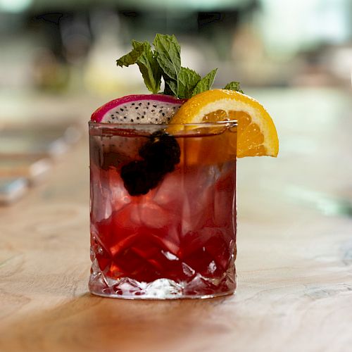 A ruby-colored cocktail in a rocks glass with ice, mint, a dragon-fruit slice, and an orange garnish.