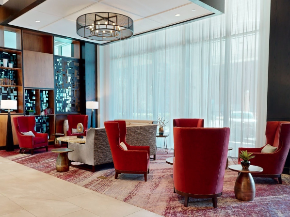 A modern hotel lounge with red armchairs, patterned rugs, glass walls, a chandelier, and a built-in bookshelf.
