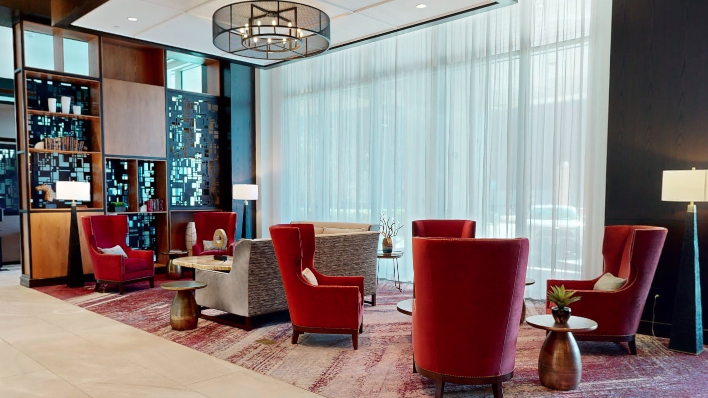 A modern hotel lounge with red armchairs, patterned rugs, glass walls, a chandelier, and a built-in bookshelf.