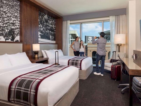 A stylish hotel room with two beds, lamps and TVs, where two people stand chatting near the window and luggage in view.