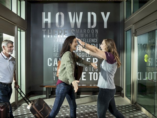 Two women and a man in an office lobby, joking and posing in front of a large &ldquo;HOWDY&rdquo; sign.