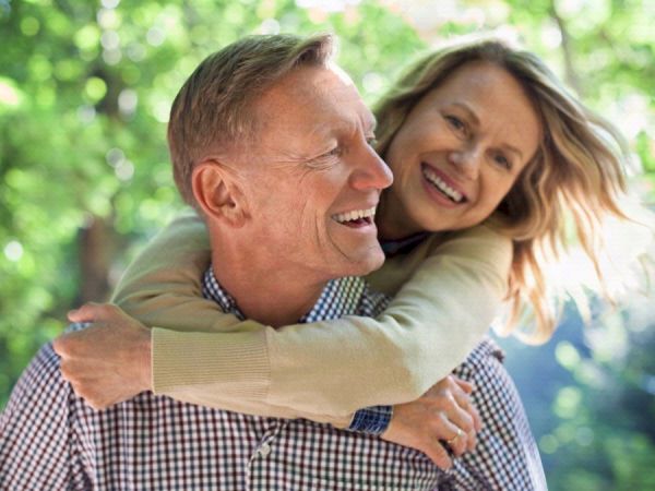 An older man smiles as a woman hugs him from behind outdoors in lush green park scenery.