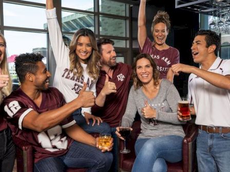A group of Texas A&M fans cheer together in a room, raising hands and holding drinks during a celebration.