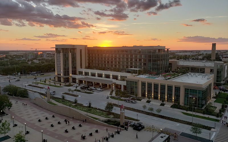 A campus building complex at sunset with dramatic clouds, cars and sidewalks below, and the sun setting on the horizon.
