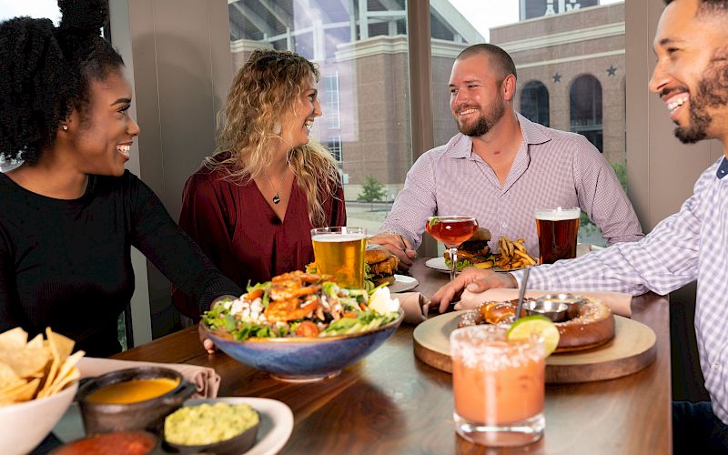 A group of friends dine outdoors at a restaurant patio, laughing over plates of food and cold drinks, together.