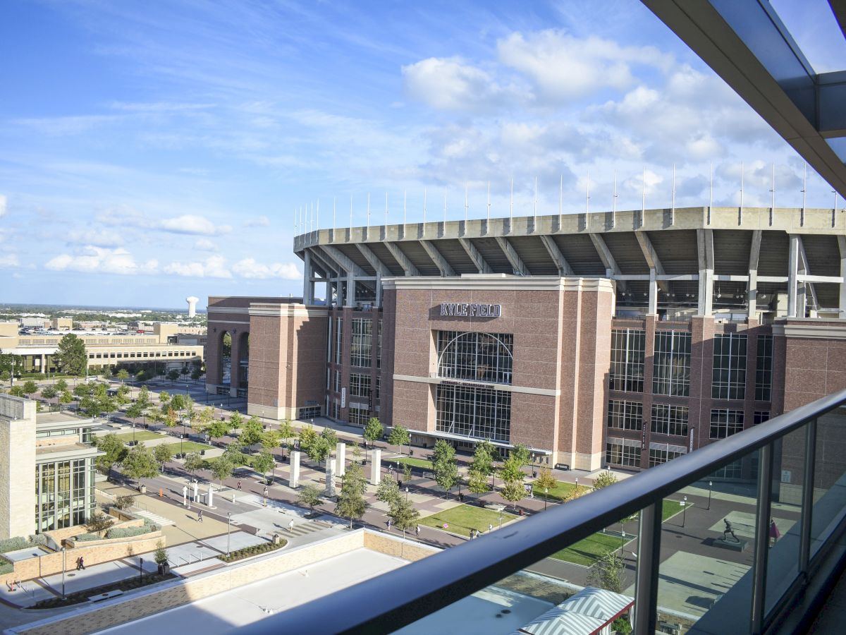A wide view of Kyle Field stadium with the &ldquo;KYLE FIELD&rdquo; sign on the brick facade and a big steel roof under blue skies.