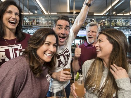A group of friends in maroon cheers at a bar, holding drinks and smiling during a game watch party.