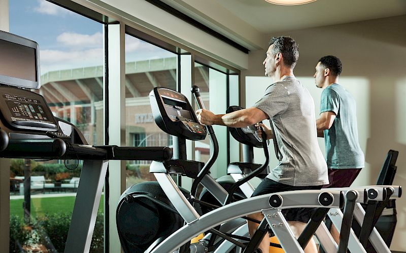 Two men work out on treadmills in a modern gym, exercising while looking out through large windows at the stadium view outside.