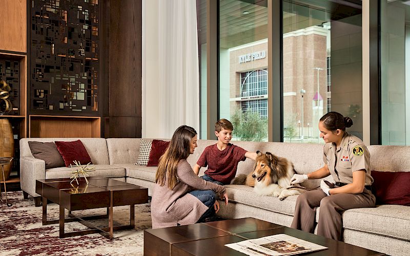 A uniformed handler and two kids pet a dog on a couch in a modern indoor lobby, while a window shows a building outside.