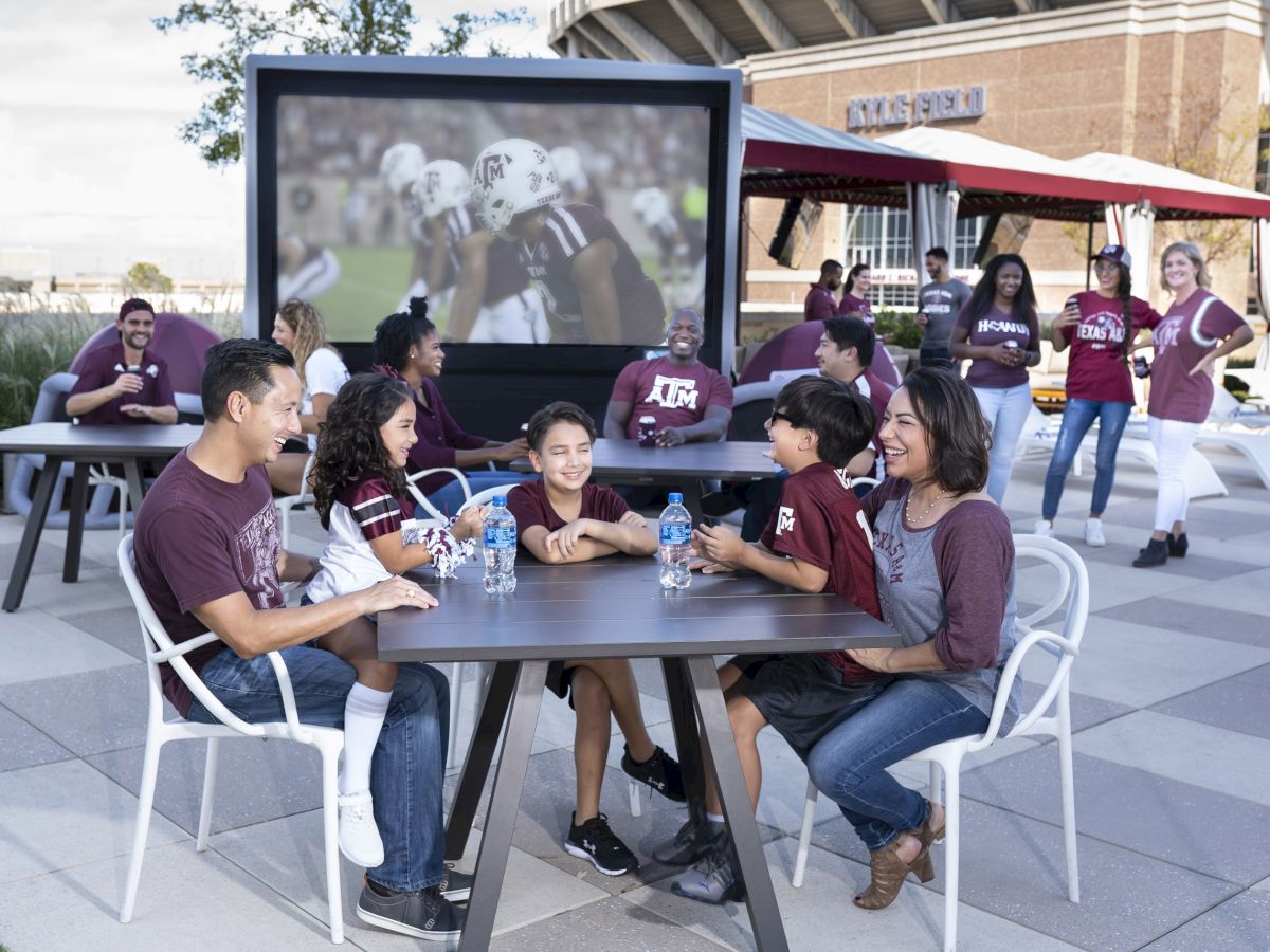 A group of people enjoying a football game on a large outdoor screen, wearing team colors, while seated at tables and socializing outdoors.