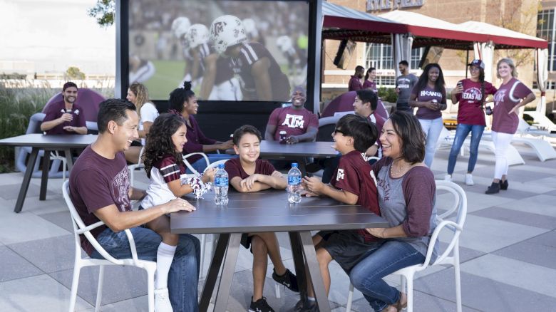 A group of people enjoying a football game on a large outdoor screen, wearing team colors, while seated at tables and socializing outdoors.