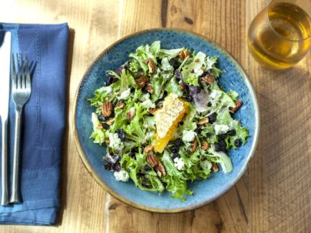 A plate of salad with mixed greens, pecans, and cheese next to a fork and knife wrapped in a blue napkin, and a glass of amber-colored drink.