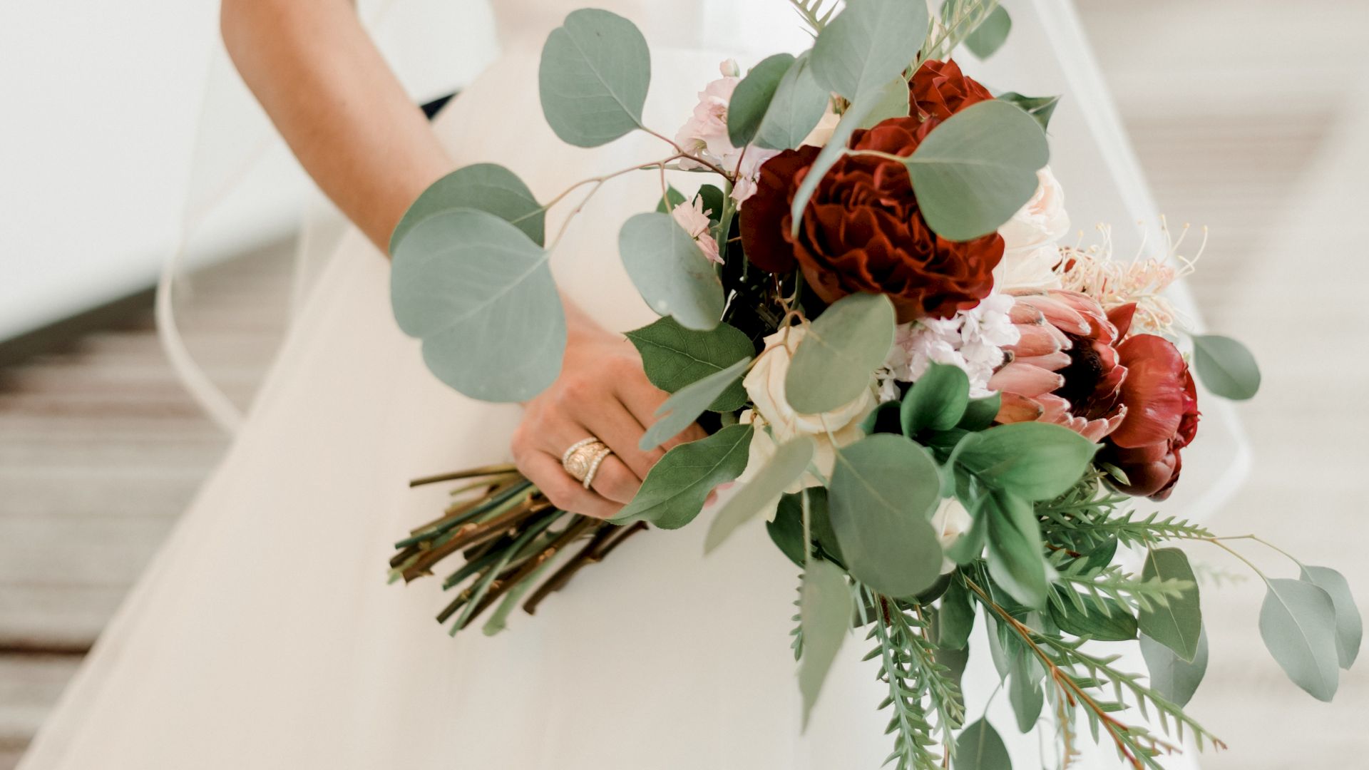 A bride in a white wedding dress holds a bouquet with green leaves and red flowers, standing in a well-lit indoor hallway.