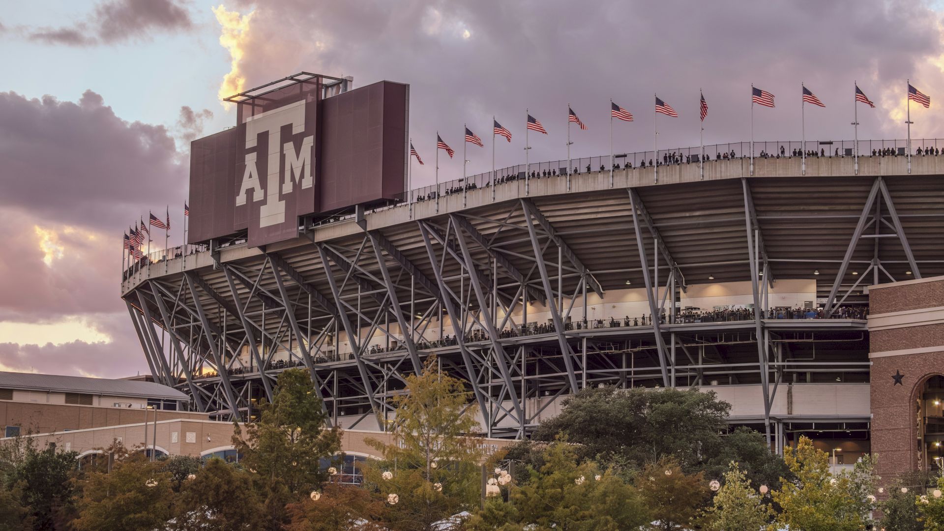 A large stadium at dusk with Texas A&M&rsquo;s &ldquo;12th Man&rdquo; on the scoreboard and many American flags waving above the stands.