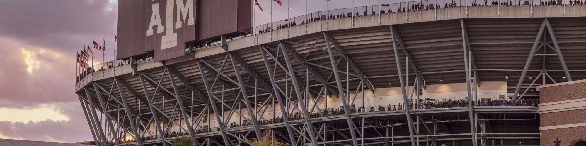 A large stadium at dusk with Texas A&M&rsquo;s &ldquo;12th Man&rdquo; on the scoreboard and many American flags waving above the stands.