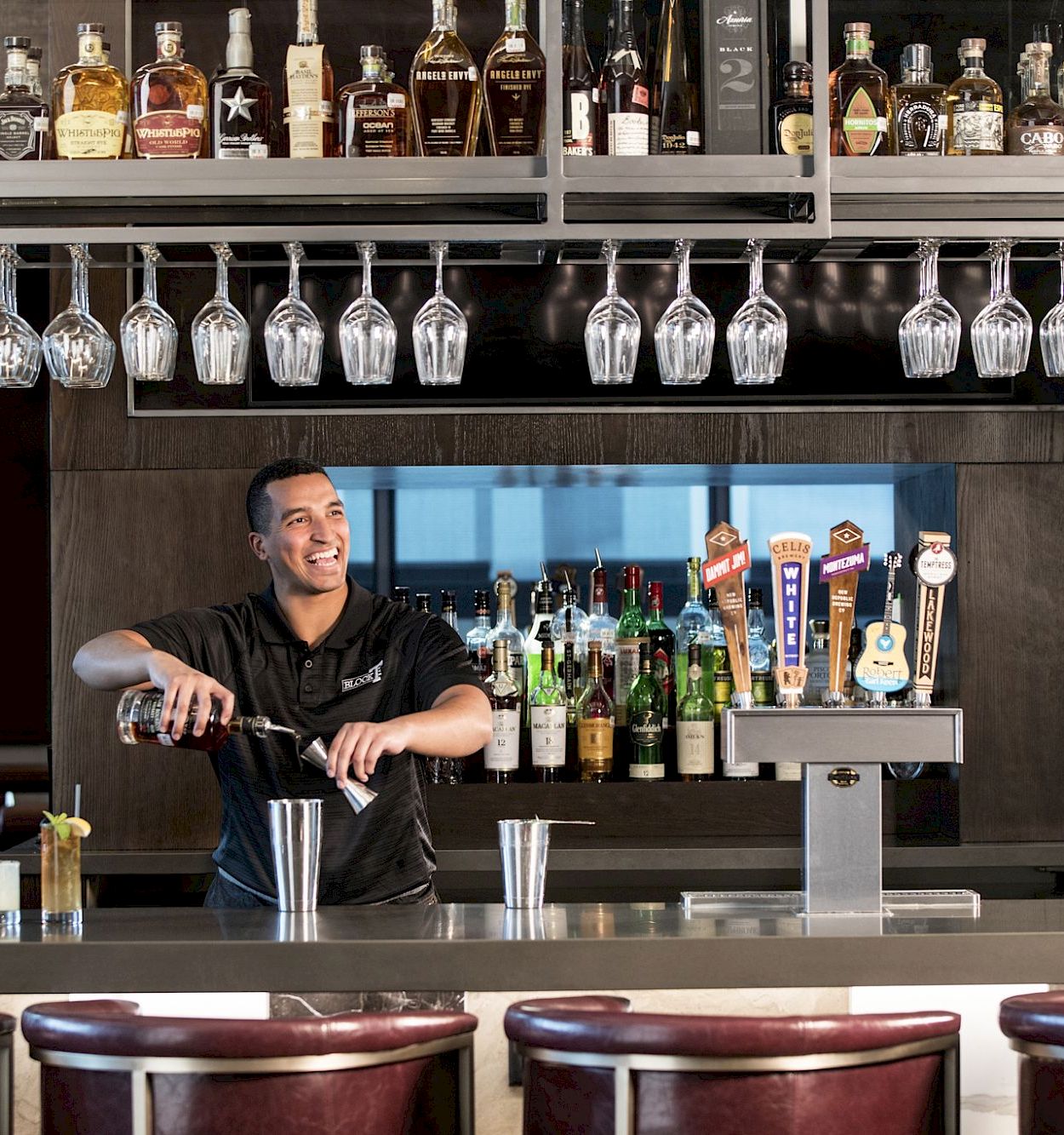 A bartender smiles behind a bar with a row of bottles and hanging glassware, preparing drinks for customers.