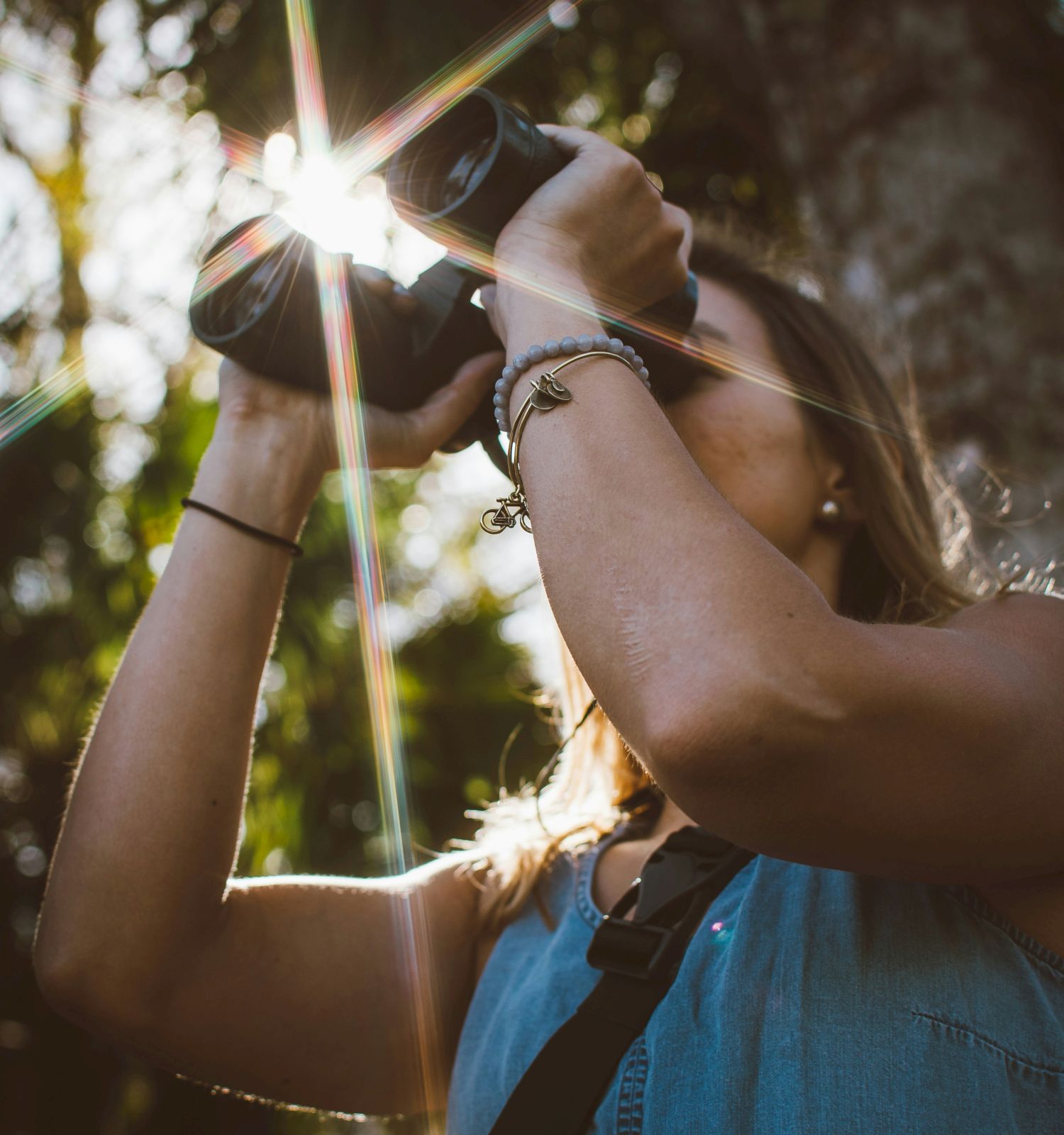 A person outdoors holds binoculars or a camera as bright sun flares through trees, creating a rainbow lens effect.
