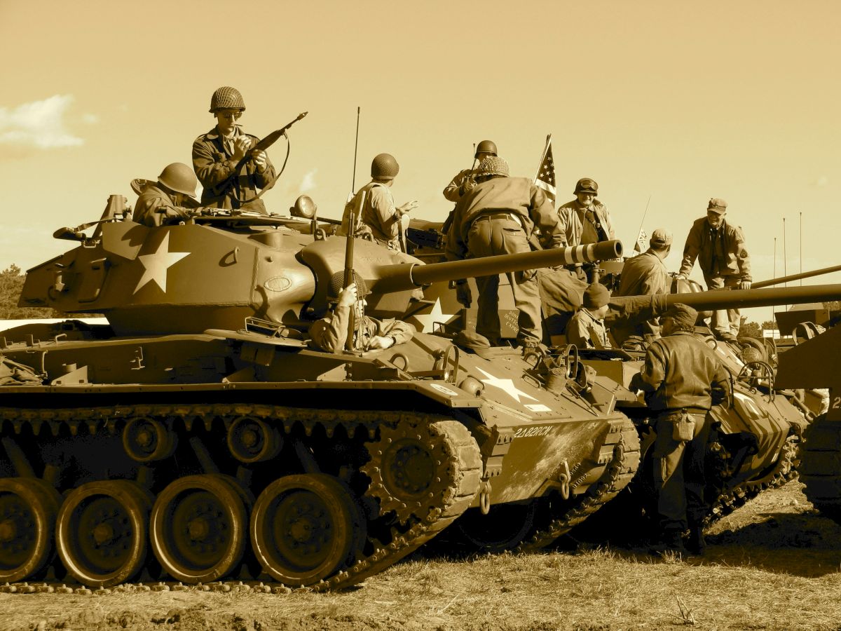 Soldiers in helmets ride an armored tank, some aiming rifles while others hold weapons, with an American flag visible.