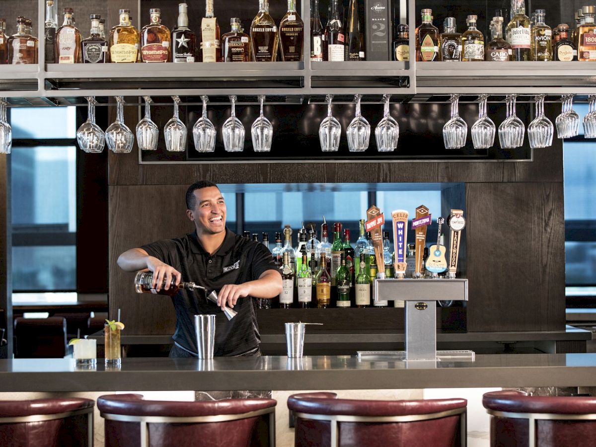 A bartender smiles while pouring at a bar stocked with liquor bottles and hanging wine glasses.