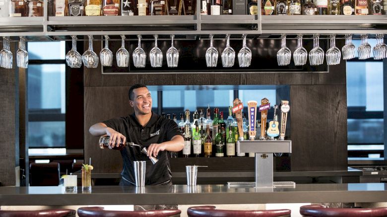 A bartender smiles while pouring at a bar stocked with liquor bottles and hanging wine glasses.
