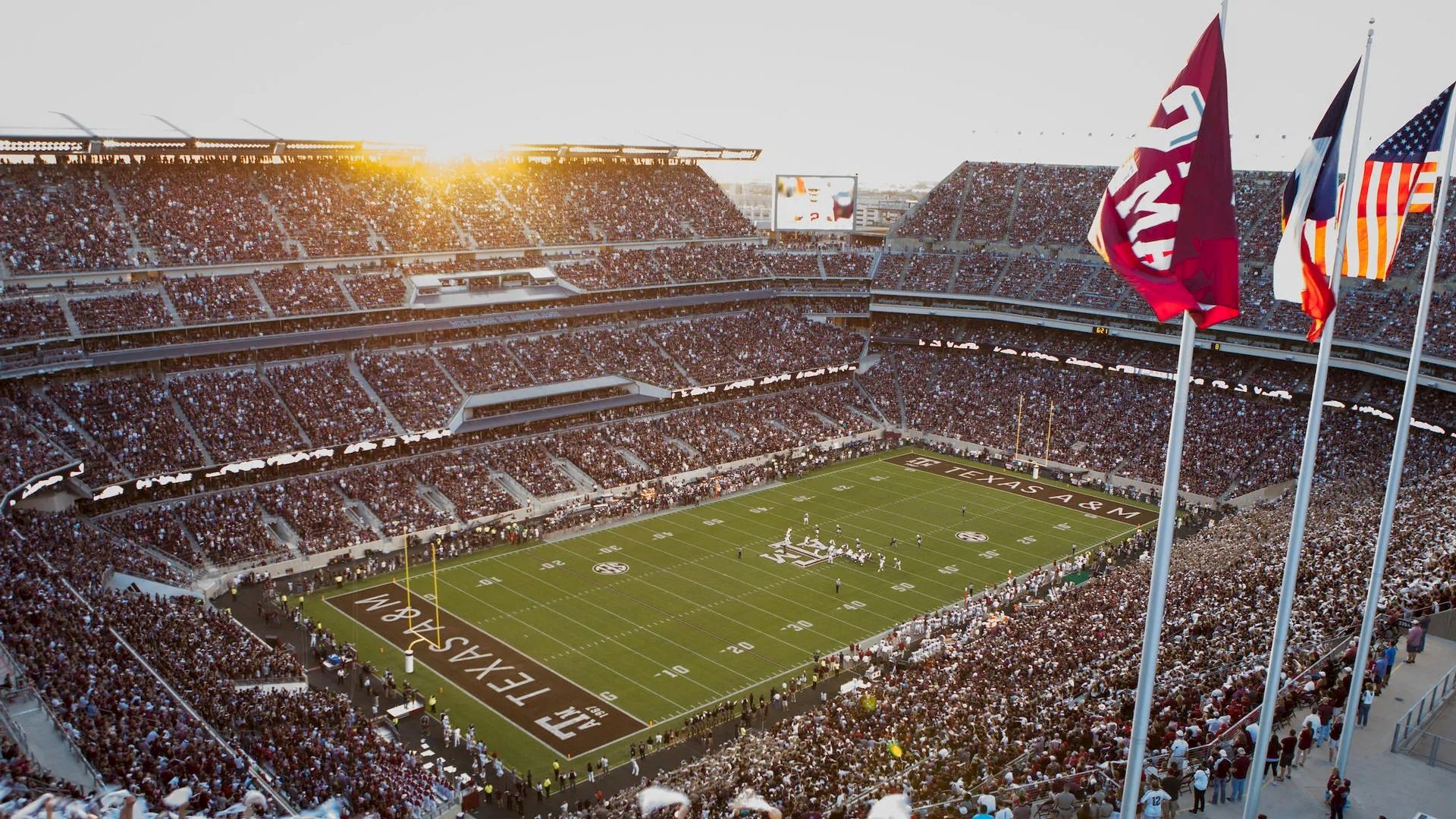 A packed football stadium during a game with "TEXAS A&M" on the field, flags waving, and the sun setting in the background ends the sentence.
